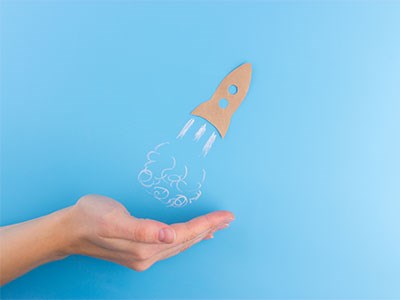 A hand beneath a sketched rocket ship taking off against a light blue background.