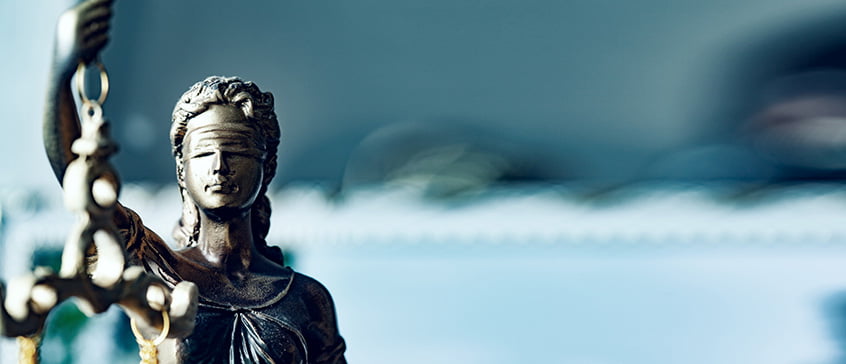 A bronze statue of Lady Justice, blindfolded and holding scales, is positioned on the left side of the frame. Her face is solemn, and the scales are slightly blurred. The background is a soft, out-of-focus blend of blue and white, suggesting a formal or institutional setting. The image evokes themes of law, fairness, and governance