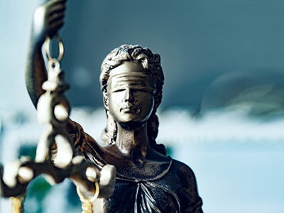 A bronze statue of Lady Justice, blindfolded and holding scales, is positioned on the left side of the frame. Her face is solemn, and the scales are slightly blurred. The background is a soft, out-of-focus blend of blue and white, suggesting a formal or institutional setting. The image evokes themes of law, fairness, and governance