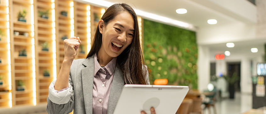 Woman in an office area looking at a tablet screen, smiling and cheering because she has good news. 