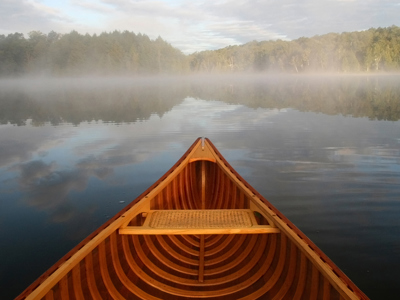 View from a canoe. The prow is visible as you look out over a calm lake to the tree-lined shore. Morning mist lightly hovers over the lake in bright morning sun.