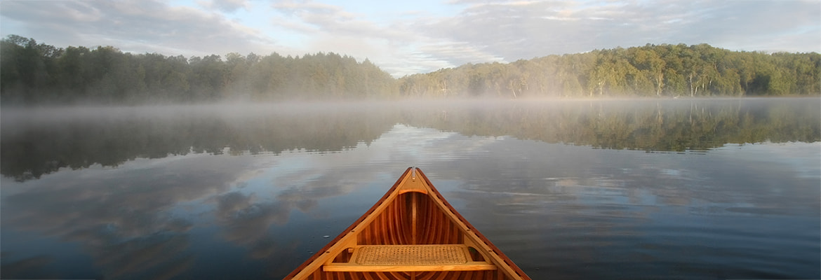 View from a canoe. The prow is visible as you look out over a calm lake to the tree-lined shore. Morning mist lightly hovers over the lake in bright morning sun.