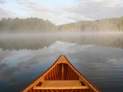 View from a canoe. The prow is visible as you look out over a calm lake to the tree-lined shore. Morning mist lightly hovers over the lake in bright morning sun.