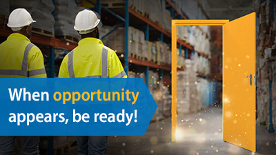 Two warehouse workers in white hard hats and high-visibility yellow safety vests stand with their backs to the camera, facing a glowing golden doorway that opens to a bright warehouse space filled with storage racks. Sparkling light particles emanate from the doorway. Overlay text reads 'When opportunity appears, be ready!' with 'opportunity' highlighted in orange.