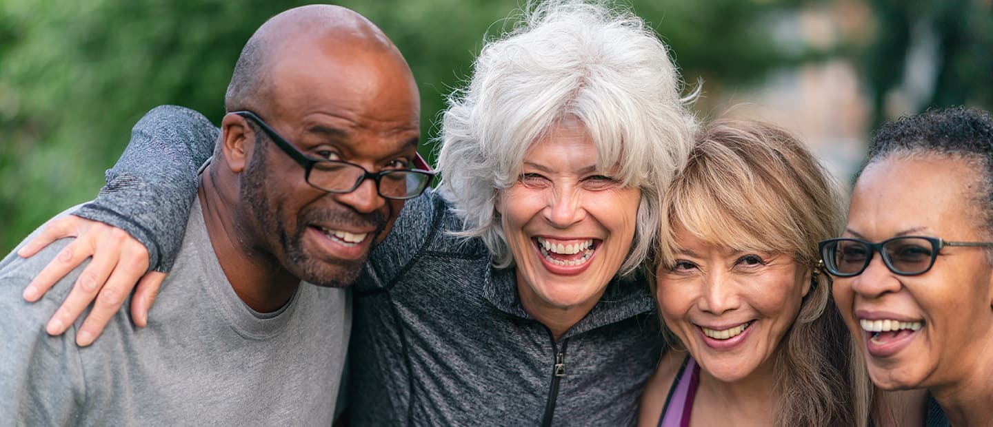 Portrait of a multi-ethnic group of senior friends at a park. The individuals are standing with arms around each others shoulders. They are smiling confidently directly at the camera. They are wearing casual athletic clothing