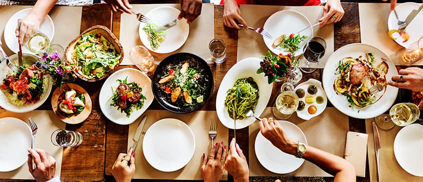 A diverse group of people gathered around a wooden table sharing a colorful, abundant meal. The table is filled with dishes like salads, pasta, roasted vegetables, and meats, emphasizing the communal experience of eating together and the cultural and environmental connections food fosters.