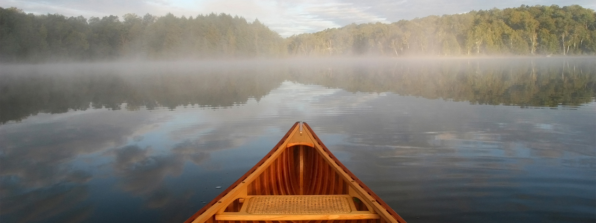 View from a canoe. The prow is visible as you look out over a calm lake to the tree-lined shore. Morning mist lightly hovers over the lake in bright morning sun.