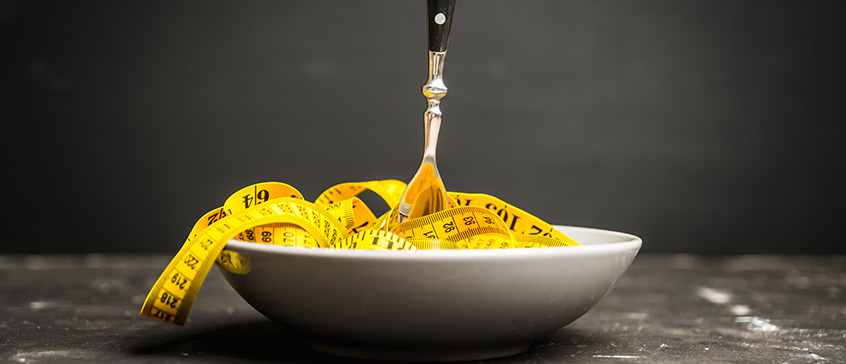 A gray ceramic bowl is filled with a coiled yellow measuring tape, with a silver fork stuck upright into the center of the tape. The measuring tape
