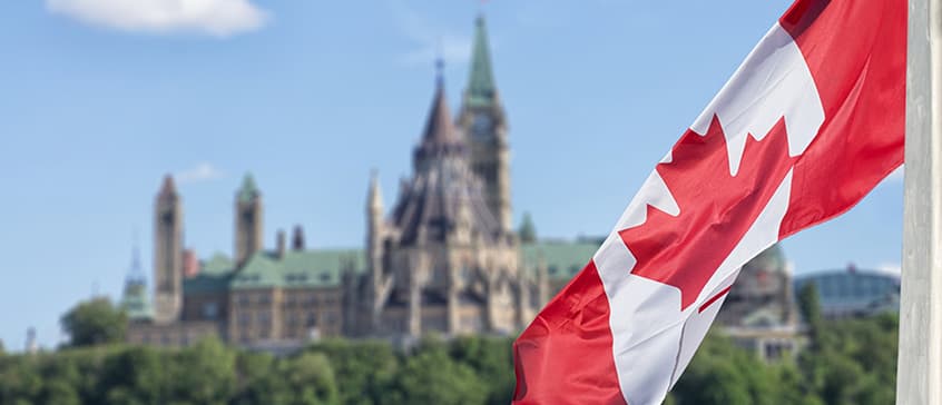 Photo of Canadian flag in the foreground and the parliament buildings in the background. 