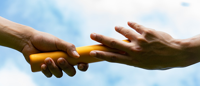 Two hands passing a golden relay baton against a blue sky with white clouds, symbolizing leadership transition and collaboration in times of change.