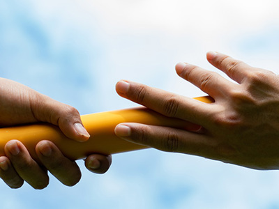 Two hands passing a golden relay baton against a blue sky with white clouds, symbolizing leadership transition and collaboration in times of change.
