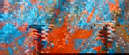 Close-up of hands holding paintbrushes creating an energetic abstract painting with vibrant orange and blue paint splatters and brushstrokes on canvas, demonstrating dynamic painting techniques and colour application.