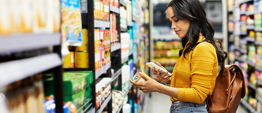 A young woman standing in a food aisle of a grocery store reading the label of a can of food and consulting information on her phone