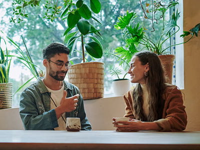 Smiling young man and woman talking over coffee together at a table in a cafe with lush, green potted plants on a wide window sill behind them.