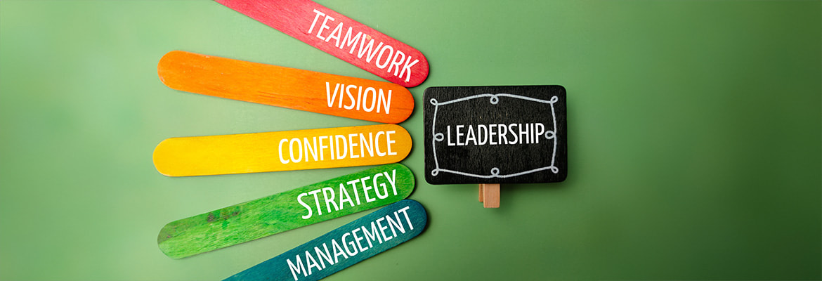 An overhead shot on a green background features five colorful popsicle sticks fanned out on the left, each with a white word: TEAMWORK (red), VISION (orange), CONFIDENCE (yellow), STRATEGY (light green), and MANAGEMENT (blue-green). To their right, a small black chalkboard sign with a white chalk border reads LEADERSHIP in white, suggesting key attributes for public relations leadership.