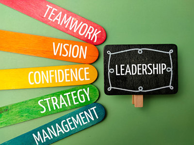 An overhead shot on a green background features five colorful popsicle sticks fanned out on the left, each with a white word: TEAMWORK (red), VISION (orange), CONFIDENCE (yellow), STRATEGY (light green), and MANAGEMENT (blue-green). To their right, a small black chalkboard sign with a white chalk border reads LEADERSHIP in white, suggesting key attributes for public relations leadership.