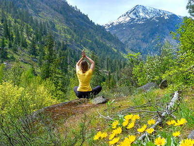 A person in a yellow top meditates on a hillside with arms raised, facing snow-capped mountains and forested valleys. Yellow wildflowers bloom in the foreground, symbolizing the connection between mindfulness practice and nature.