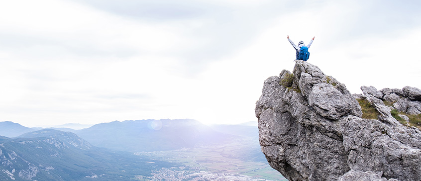 Hiker with backpack standing triumphantly atop a rocky mountain summit with arms raised in victory, overlooking a vast valley and mountain ranges below under an overcast sky, symbolizing overcoming challenges and reaching new heights.