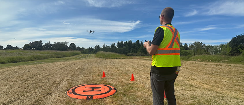 A person in a neon yellow safety vest with orange stripes operates a drone in an open grassy field. The drone is flying above, and an orange landing pad with an "H" symbol is on the ground, flanked by two small orange cones. Trees and a blue sky with scattered clouds are in the background.