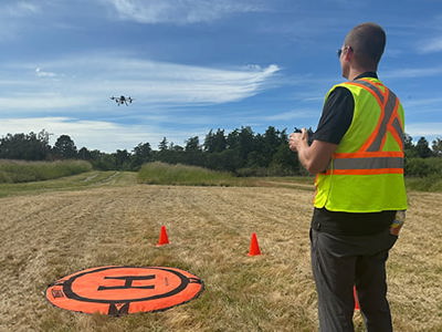 A person in a neon yellow safety vest with orange stripes operates a drone in an open grassy field. The drone is flying above, and an orange landing pad with an "H" symbol is on the ground, flanked by two small orange cones. Trees and a blue sky with scattered clouds are in the background.
