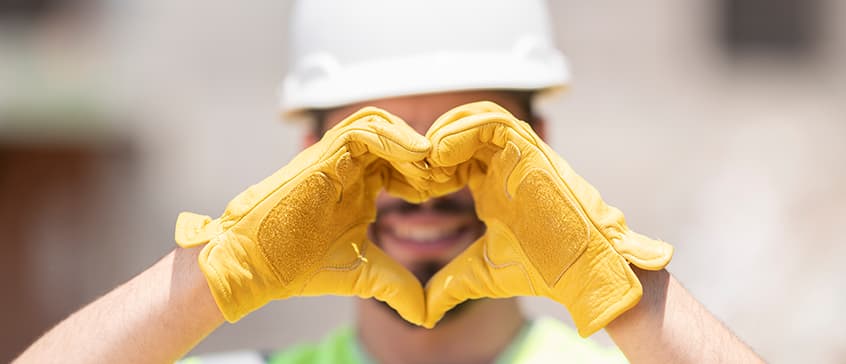 Man facing forward wearing a white hardhat and yellow leather work gloves makes a heart sign with his hands in front of his face