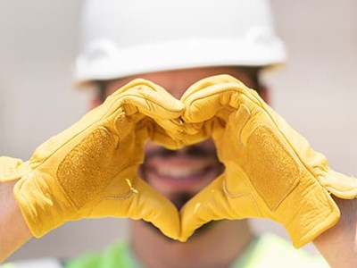 Man facing forward wearing a white hardhat and yellow leather work gloves makes a heart sign with his hands in front of his face
