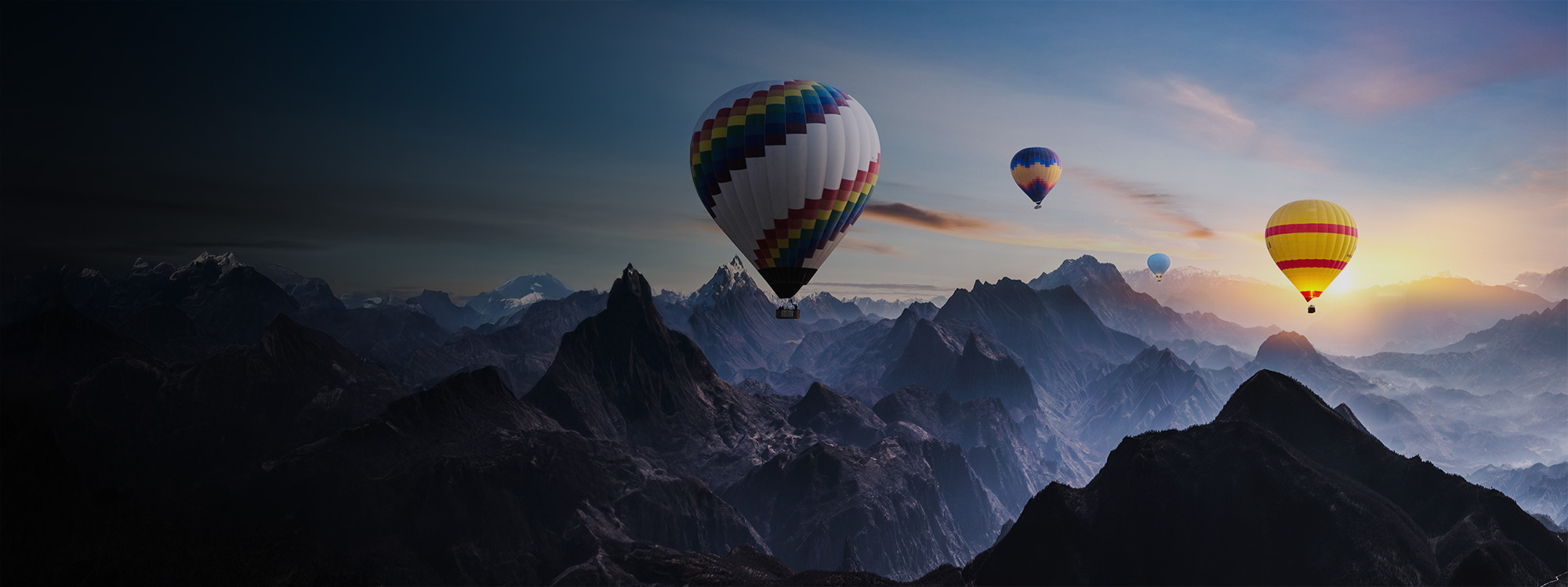 Four colourful hot air balloons float above a dramatic mountain range at sunrise, with soft light illuminating the peaks and sky.