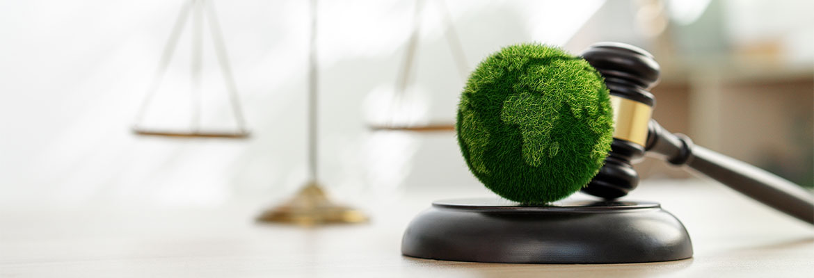  A gavel rests beside a green globe covered in grass, with a justice scale in the background.