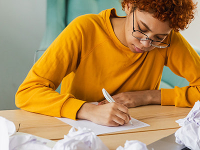 A person with curly red hair and glasses wearing a yellow sweater sits at a wooden desk, focused on writing by hand on paper. Crumpled papers are scattered around the workspace, with a laptop open nearby, illustrating the creative writing process and revision work.