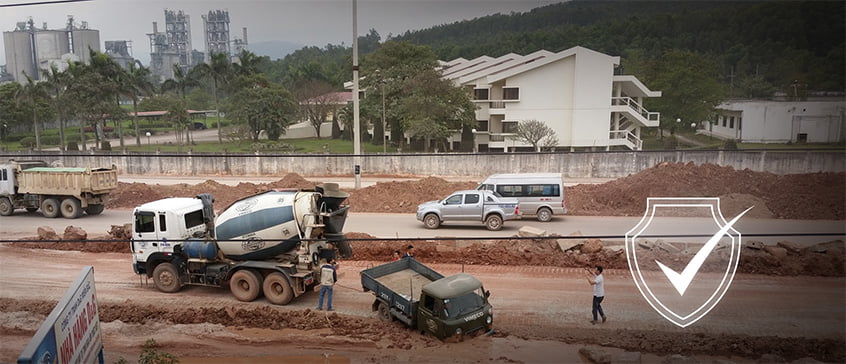 Construction site with concrete mixer trucks and dump trucks on dusty, unpaved ground. Workers visible near heavy machinery without clear protective equipment. Residential buildings nearby suggest potential community exposure to construction dust and noise pollution, impacting both worker respiratory health and air quality for local residents.