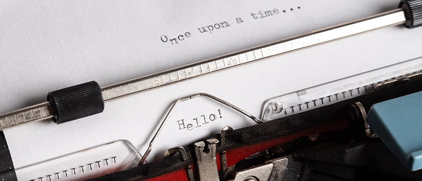 A close-up shot of an old-fashioned typewriter with a piece of white paper inserted. At the top of the visible paper, the phrase "Once upon a time..." is typed. Further down, in the center, the word "Hello!" is typed. The metal components of the typewriter, including the platen and type guide, are in sharp focus. The image evokes the act of writing, self-reflection, and the beginning of a narrative