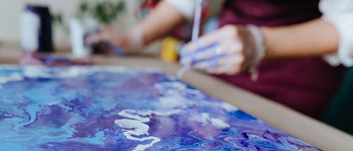 Close-up of fluid acrylic paint in vibrant purple and blue tones spreading organically across canvas, with artist's hands visible working in the background, showcasing the flowing nature of pour painting techniques
