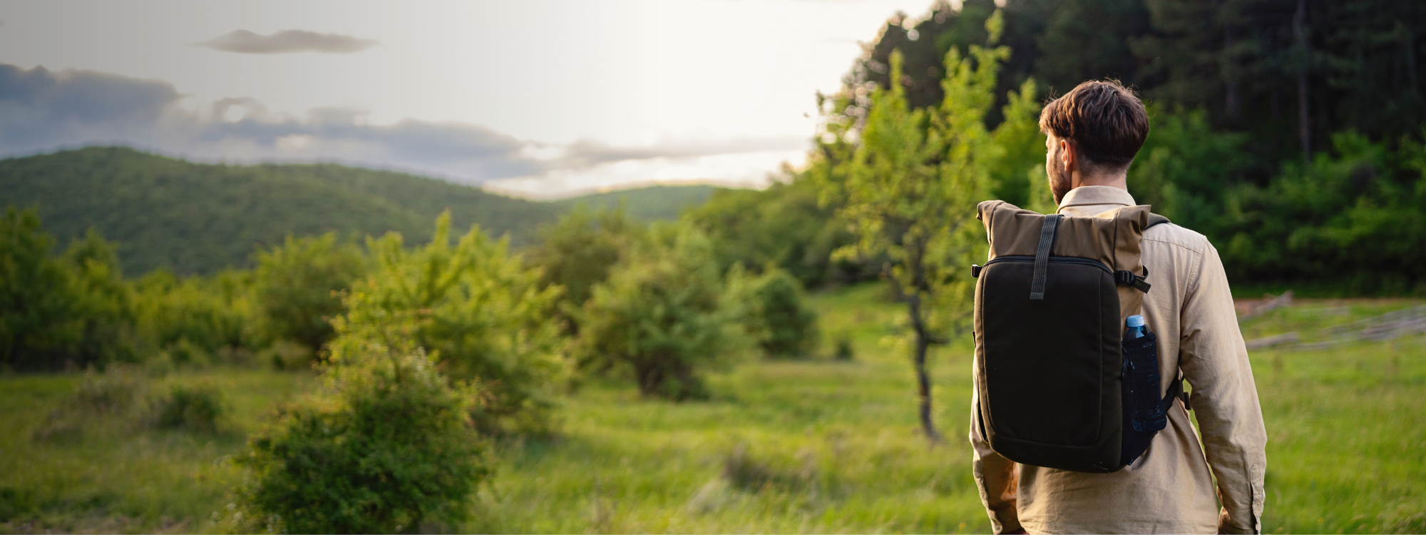 A young man stands in a grassy field, wearing a backpack, admiring the natural scenery.