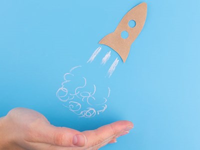 A hand beneath a sketched rocket ship taking off against a light blue background.