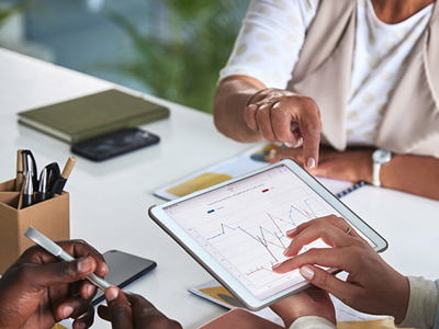 Three people collaborate around a white desk in a modern office setting, with one person pointing to data charts and graphs displayed on a tablet screen. The scene includes office supplies, a smartphone, and notebook, illustrating teamwork in data analysis and collaborative decision-making.