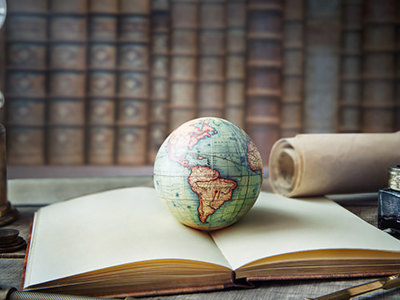Vintage study scene with an open book, a small globe, an ink bottle with a quill, rolled parchment, a magnifying glass and an oil lamp on a wooden desk, set against shelves of old books, symbolizing history and global exploration.