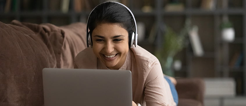 Close up smiling Indian woman in headphones using laptop, lying on couch, having fun talking with other English language students in the UVic ELC Conversation Lounge. 