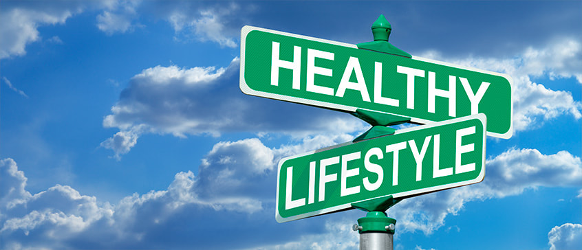 A street signpost with two green directional signs against a bright blue sky with white clouds. The top sign points left and reads HEALTHY in white capital letters. The bottom sign points right and reads LIFESTYLE in white capital letters. The image symbolizes choices and directions toward a healthier life