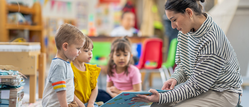An elementary school teacher sits on the floor of her classroom with a small group of students as she reads them a book. 