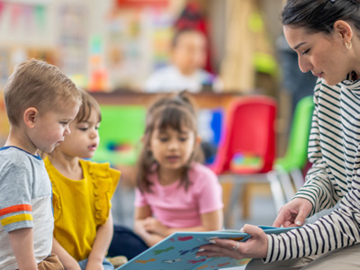 An elementary school teacher sits on the floor of her classroom with a small group of students as she reads them a book. 