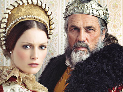 A woman and a man dressed in elaborate historical costumes pose against a patterned wallpaper background; the woman wears a gold headdress and lace gown, while the man wears a fur-trimmed robe and silver crown, evoking a royal or noble setting.