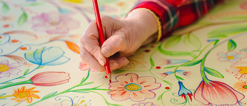 A close-up of a hand using a red coloured pencil to draw and colour detailed floral patterns on a large sheet of paper. The artwork features vibrant flowers, leaves, and stems in shades of red, blue, green, yellow and pink. The person is wearing a red plaid shirt with gold trim on the sleeve