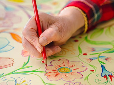 A close-up of a hand using a red coloured pencil to draw and colour detailed floral patterns on a large sheet of paper. The artwork features vibrant flowers, leaves, and stems in shades of red, blue, green, yellow and pink. The person is wearing a red plaid shirt with gold trim on the sleeve