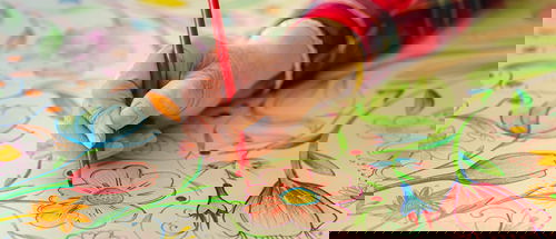 A close-up of a hand using a red coloured pencil to draw and colour detailed floral patterns on a large sheet of paper. The artwork features vibrant flowers, leaves, and stems in shades of red, blue, green, yellow and pink. The person is wearing a red plaid shirt with gold trim on the sleeve