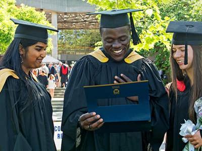 3 students on graduation day in cap and gown smiling and looking at their graduation parchments