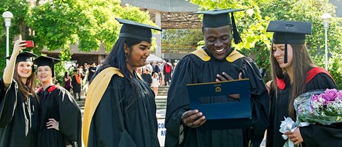 3 students on graduation day in cap and gown smiling and looking at their graduation parchments