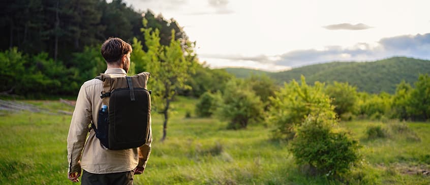 A person with a backpack walking through a lush green meadow surrounded by small trees, with forested hills and a bright sky in the background, symbolizing a journey into nature and sustainability.
