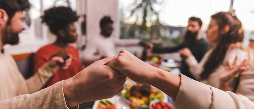 A close-up shot focuses on clasped hands of diverse individuals around a dining table, suggesting a moment of shared ritual or prayer. The soft lighting and blurred background with a meal emphasize a sense of community and connection, central to the themes of ritual, religion, and culture.