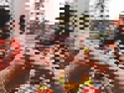 A close-up shot focuses on clasped hands of diverse individuals around a dining table, suggesting a moment of shared ritual or prayer. The soft lighting and blurred background with a meal emphasize a sense of community and connection, central to the themes of ritual, religion, and culture.