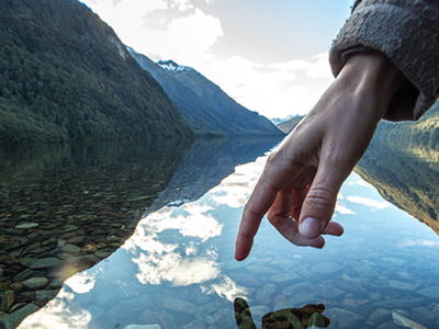 A hand gently touching the surface of clear lake water, creating small ripples. The water reflects the sky, mountains, and a few clouds. The lakebed is visible through the clear water, covered with small rocks. Green mountains rise on either side of the lake under a bright sky.